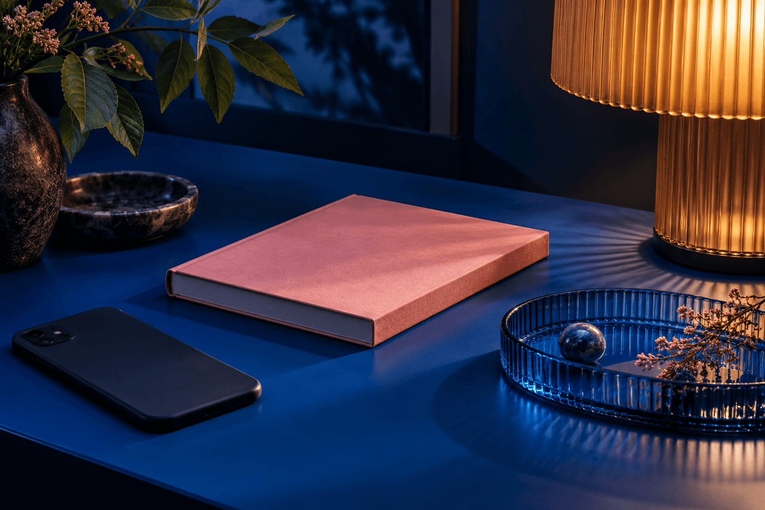 Facedown phone, blank pink book and translucent blue tray in lamplight
